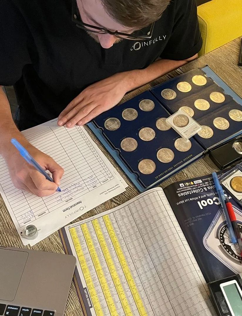 A person writes on a form while surrounded by coins, a laptop, and a reference book on coin grading.