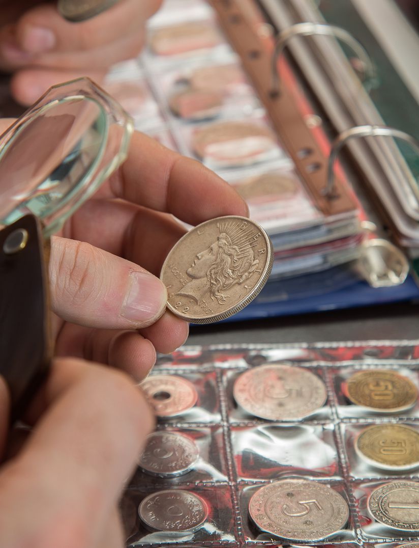 A person examines a coin with a magnifying glass, surrounded by a binder of various coins.