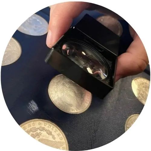 A hand holding a magnifying glass over a collection of coins on a dark surface.