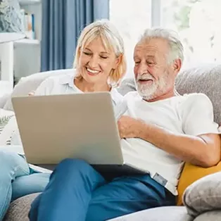 A woman and an older man smile while looking at a laptop together on a couch