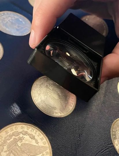 A hand holding a magnifying glass over several coins on a dark surface.