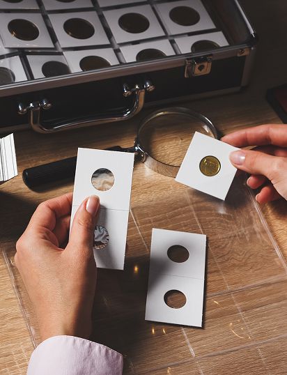 Hands holding coin holders with coins, a magnifying glass, and a coin storage case
