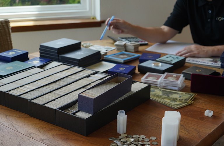 A person appraising coins on a table filled with coin holders, currency, and appraisal tools.